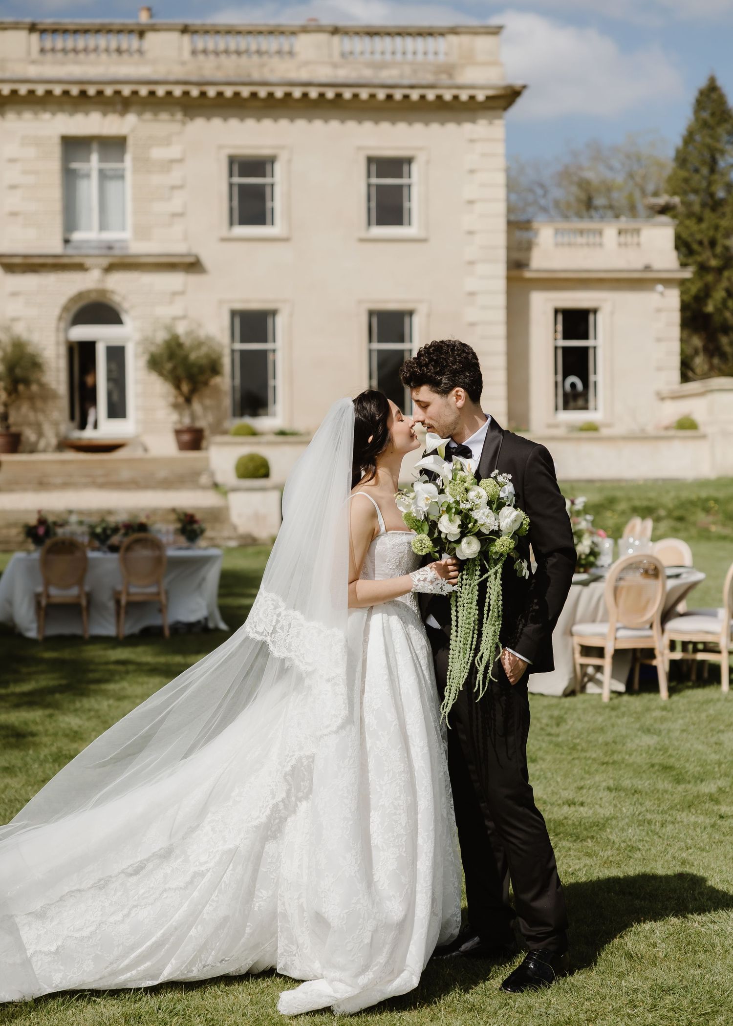 Bride and groom share a romantic moment on a lawn outside an elegant stone manor house, bride holding white bouquet.