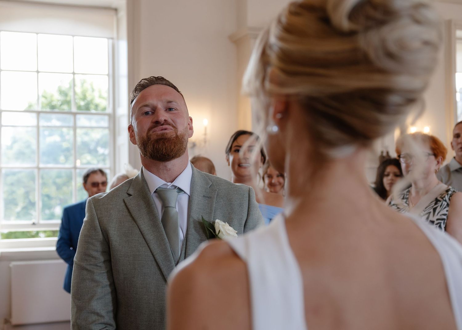 Groom in grey suit smiling emotionally at bride during indoor wedding ceremony with guests in background.