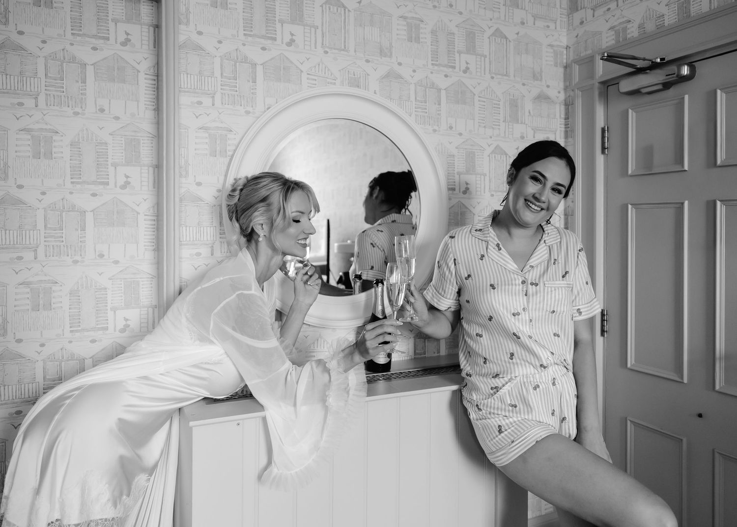 Bride in white robe and bridesmaid in pajamas toast with champagne glasses at a vanity mirror in black and white.