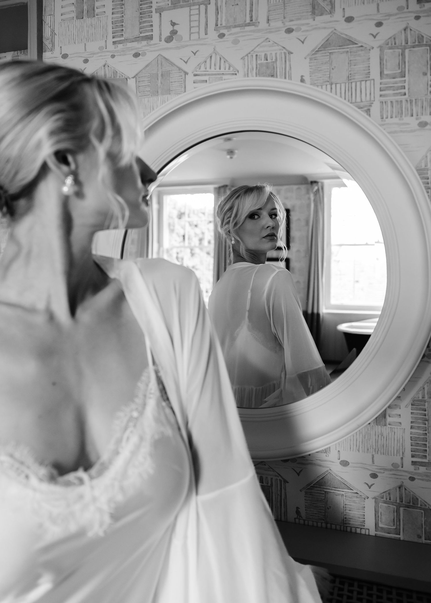 Bride in lace wedding dress and robe gazes at her reflection in a round mirror, captured in elegant black and white.