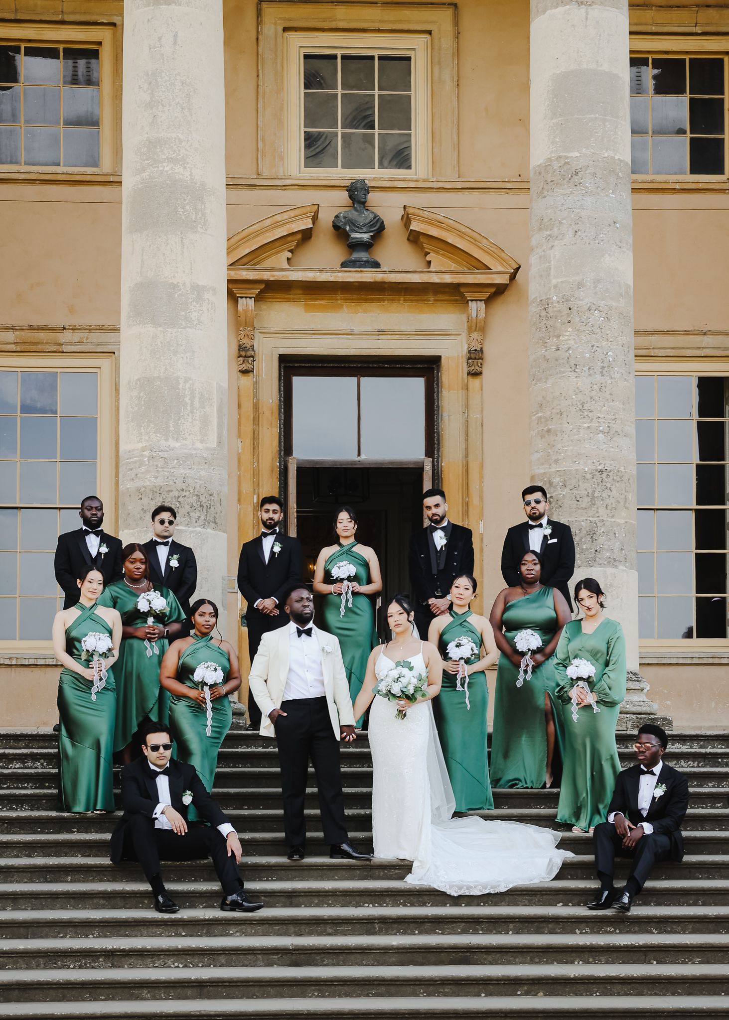 Wedding party poses on grand steps, bridesmaids in green satin gowns with groomsmen in black tuxedos flanking bride and groom.