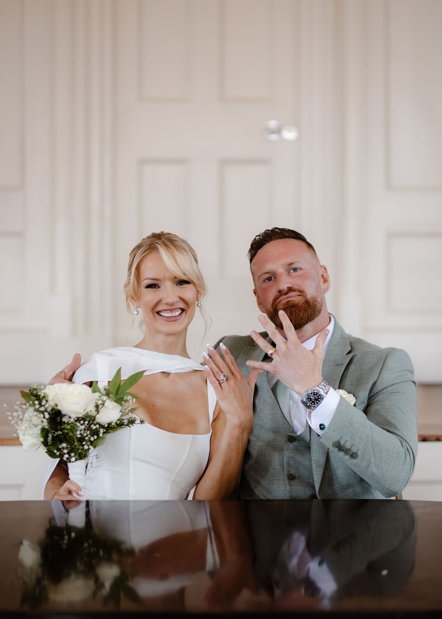 Bride and groom sitting at table showing wedding rings, bride holding white bouquet in elegant white venue.