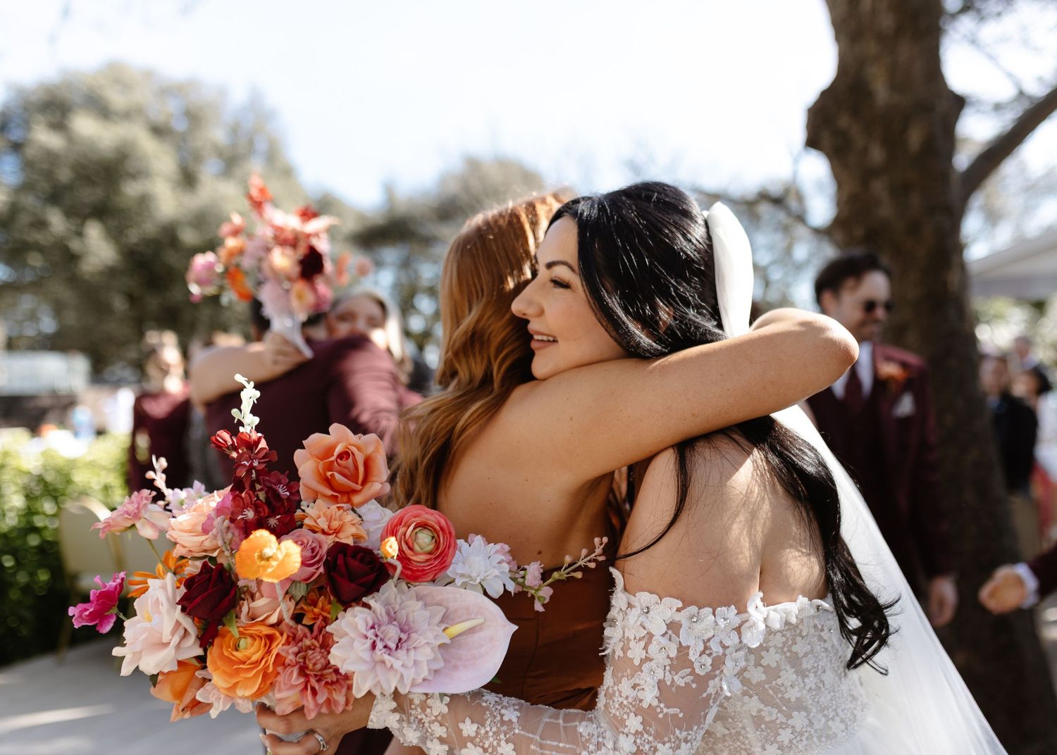 Two brides share a heartfelt hug at their wedding, one holding a vibrant bouquet of pink, red, and orange flowers.