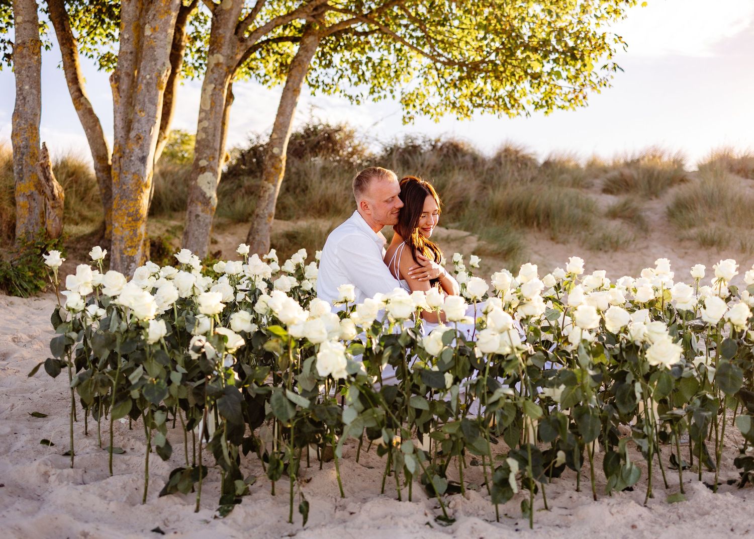Couple sitting together on sandy beach surrounded by white roses with trees in background at golden hour.