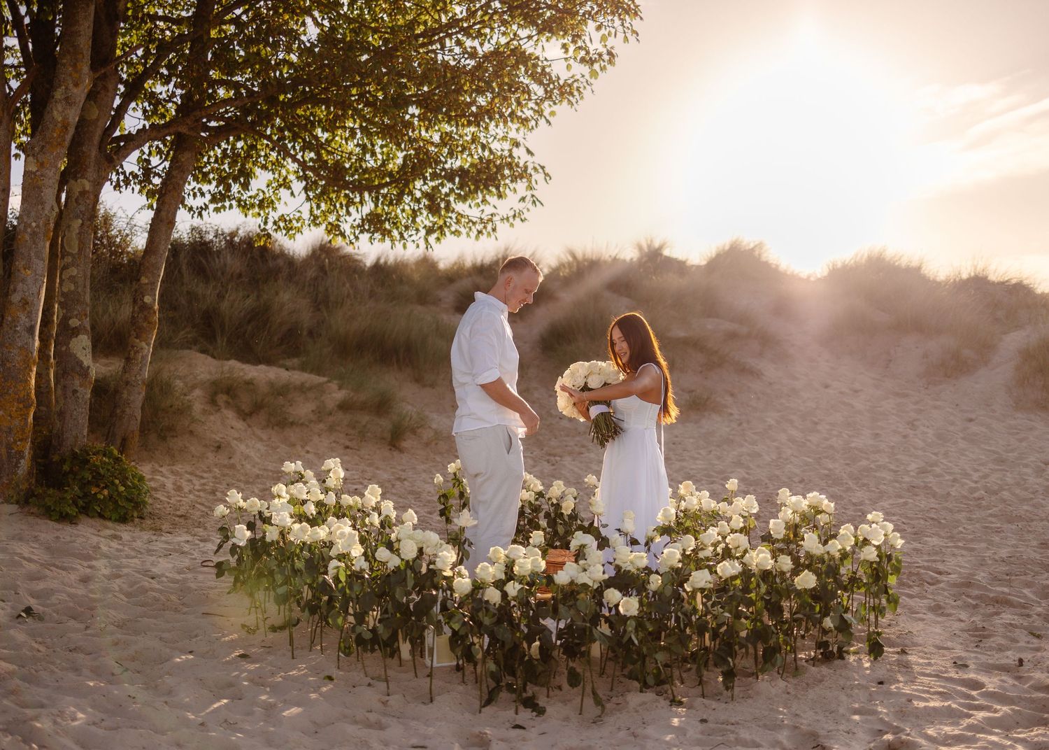 Couple in white attire standing among white floral arrangements on sandy beach dunes at golden sunset.