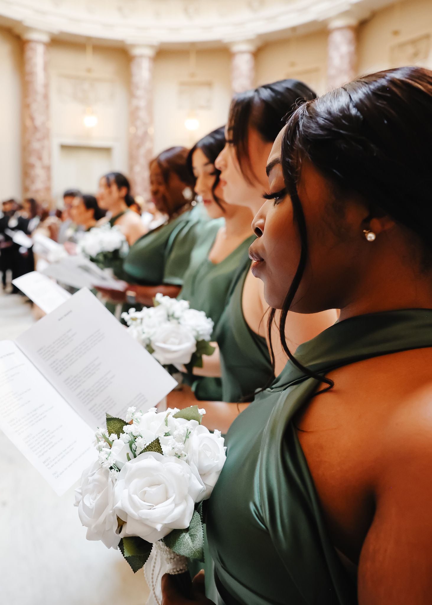 Wedding party members in sage green dresses holding white rose bouquets during ceremony.