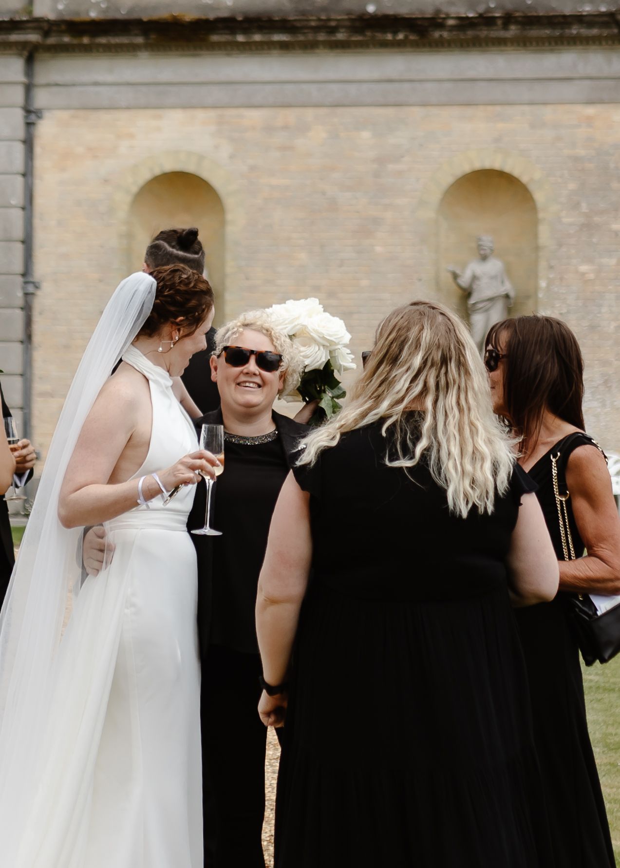 Wedding party members in black attire mingling during outdoor reception against stone building backdrop.
