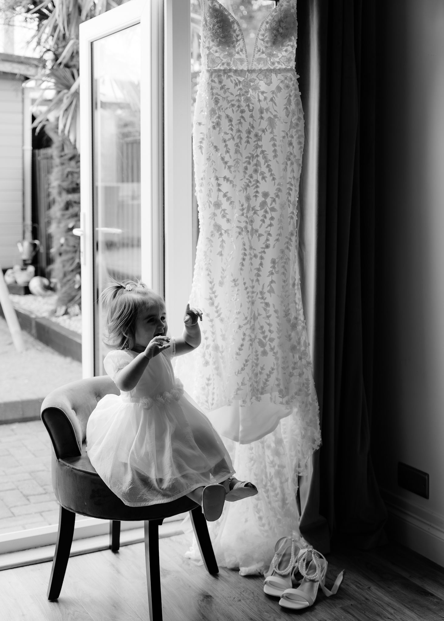 A white wedding dress and bridal shoes displayed on an elegant black chair near a window.
