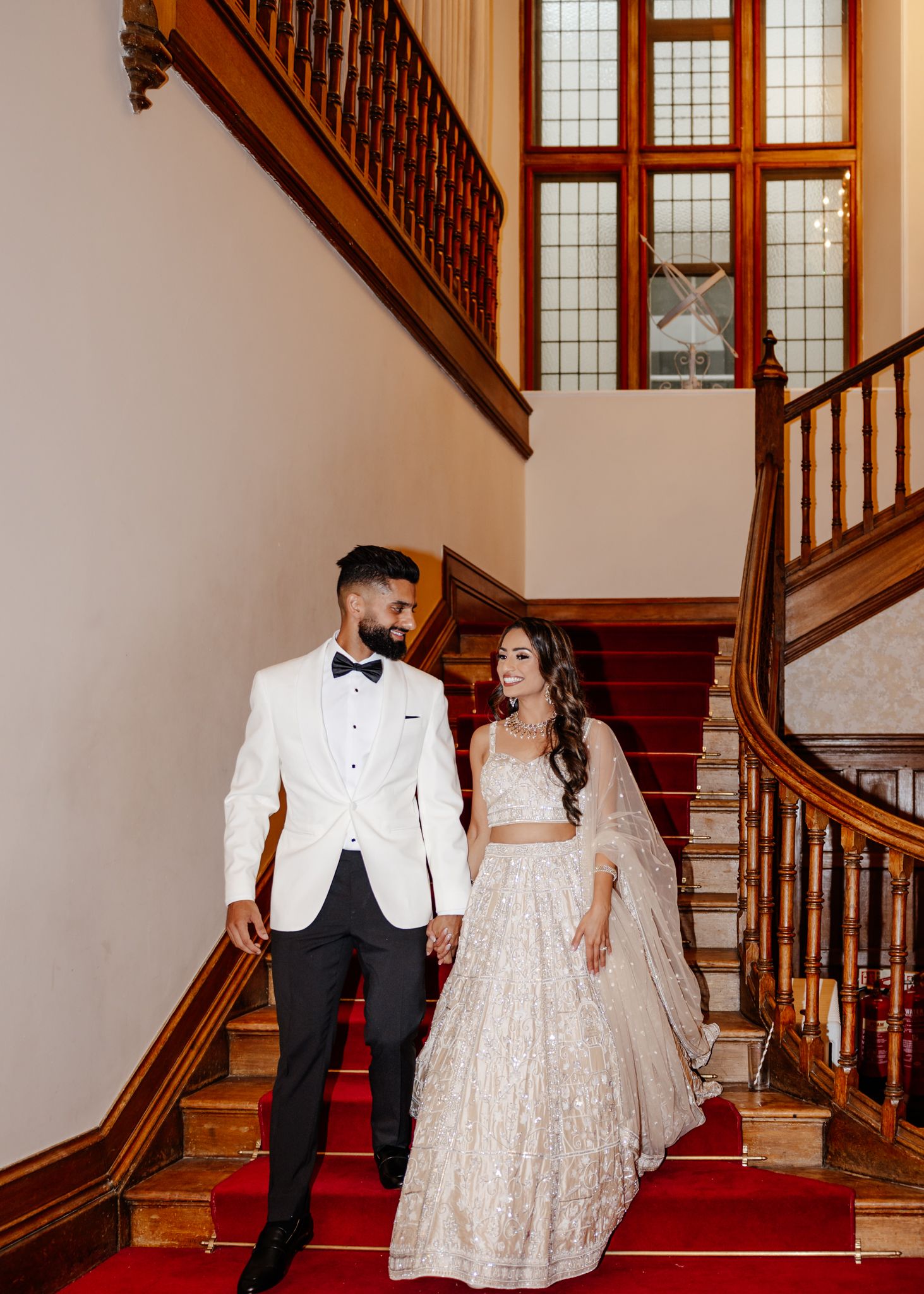 A couple descends an elegant wooden staircase with stained glass windows in formal wedding attire.