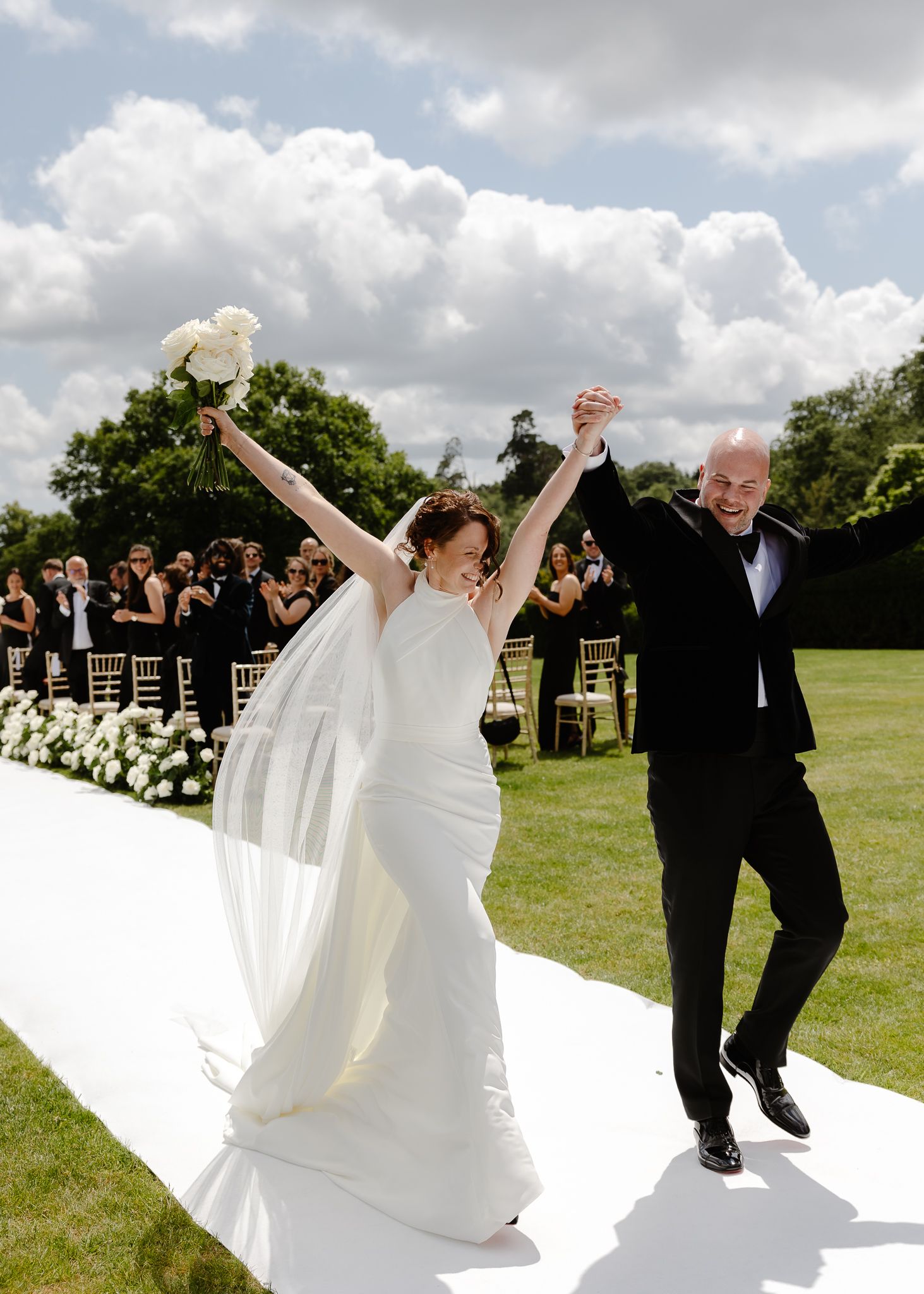 Wedding party members dance joyfully in a line across a grassy field under cloudy skies.