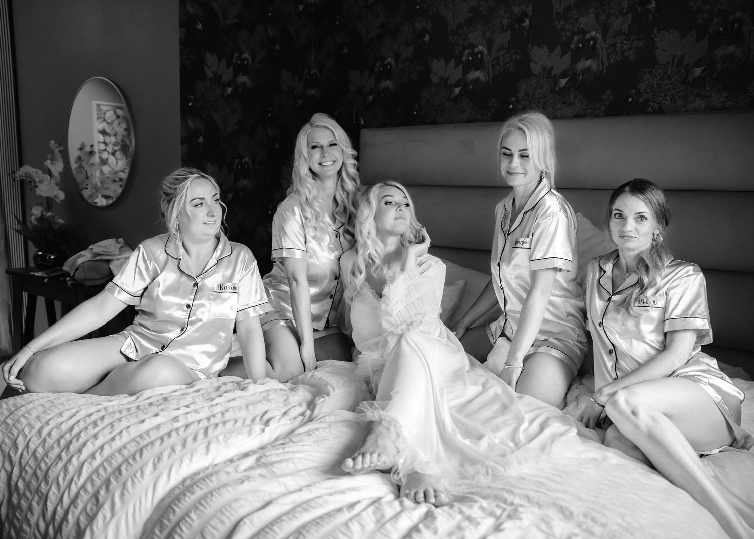 Black and white photo of a bride in white robe with four bridesmaids in satin pajamas sitting on a bed.