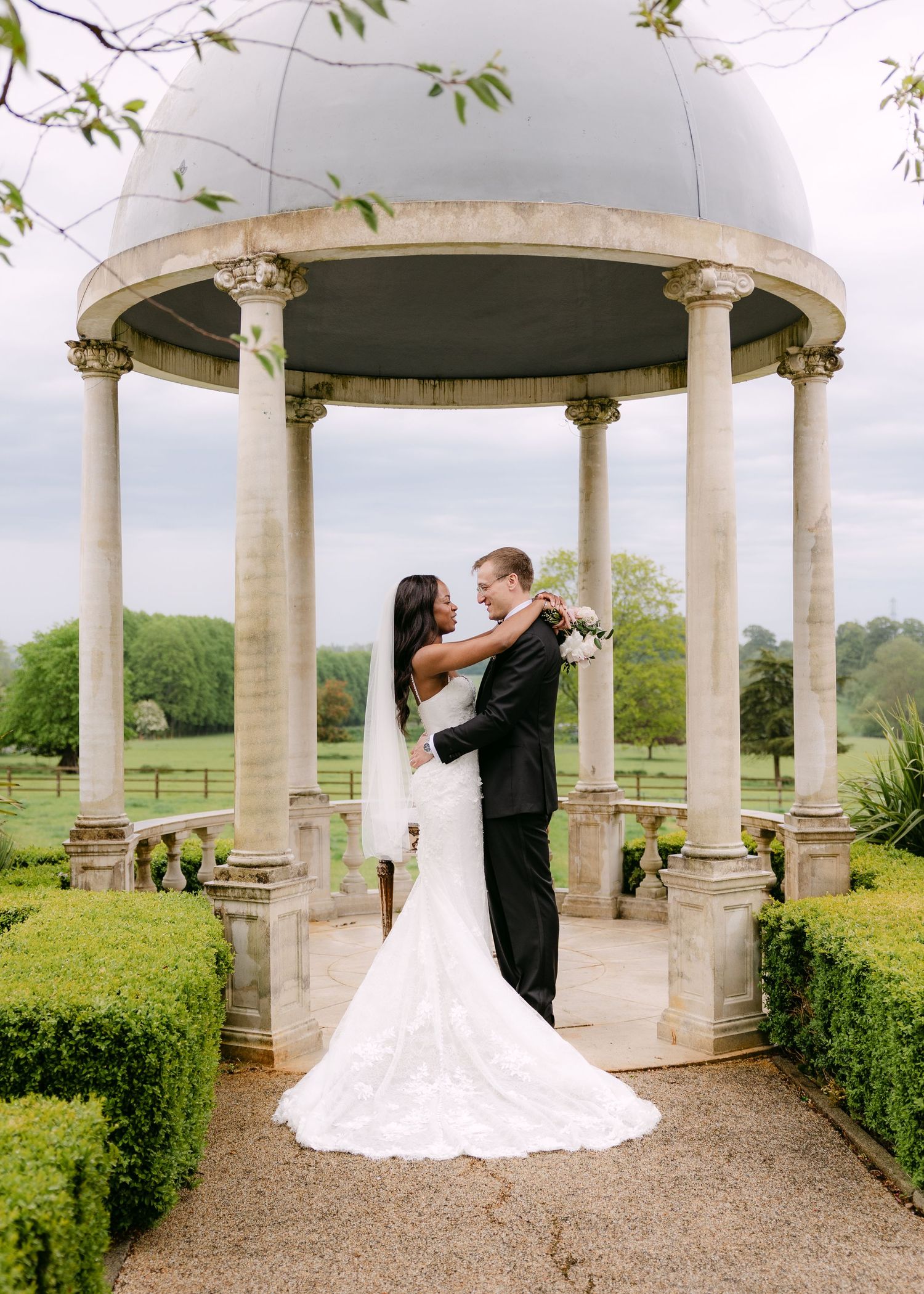 Bride and groom embrace under elegant stone gazebo with dome roof, surrounded by manicured gardens and green countryside.