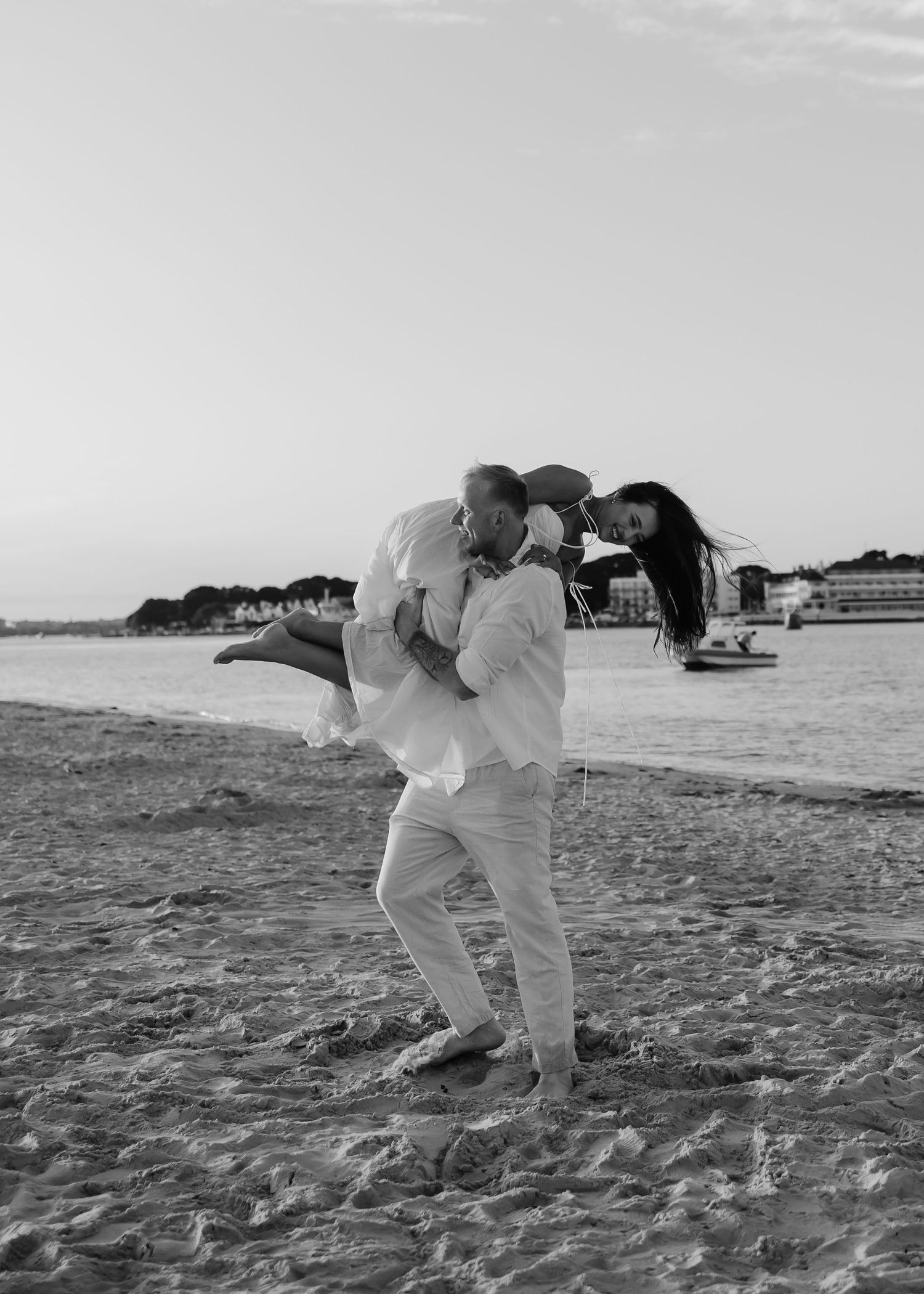 Black and white photo of a couple kissing on a beach, man lifting woman in his arms at sunset.