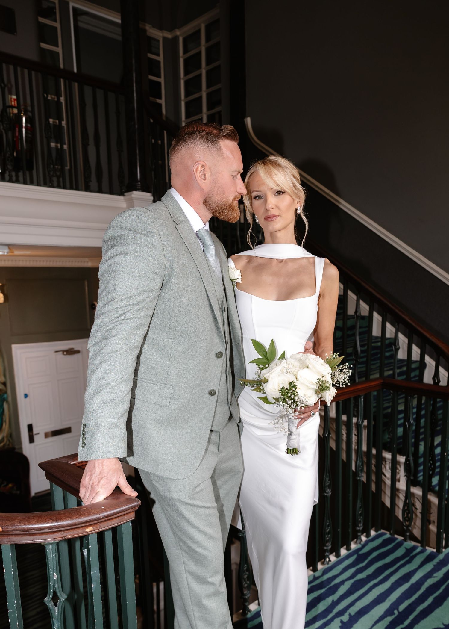 Groom in sage green suit kisses bride in sleek white gown holding white bouquet on staircase.