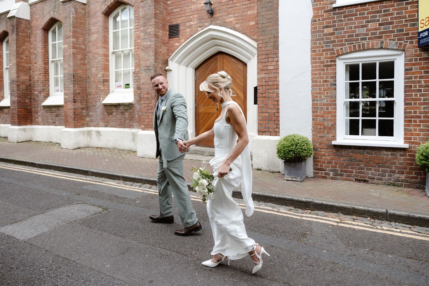 Bride and groom holding hands crossing a street outside a brick church, bride wearing white gown holding bouquet.