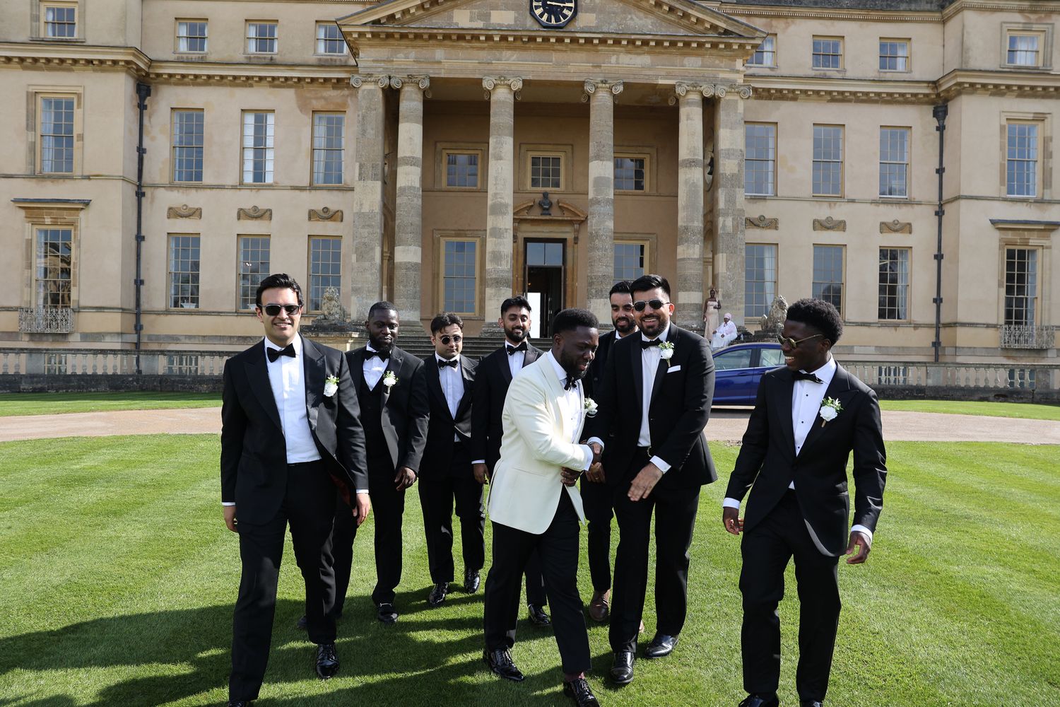 Groomsmen in black tuxedos gather on a manicured lawn in front of an elegant historic mansion.