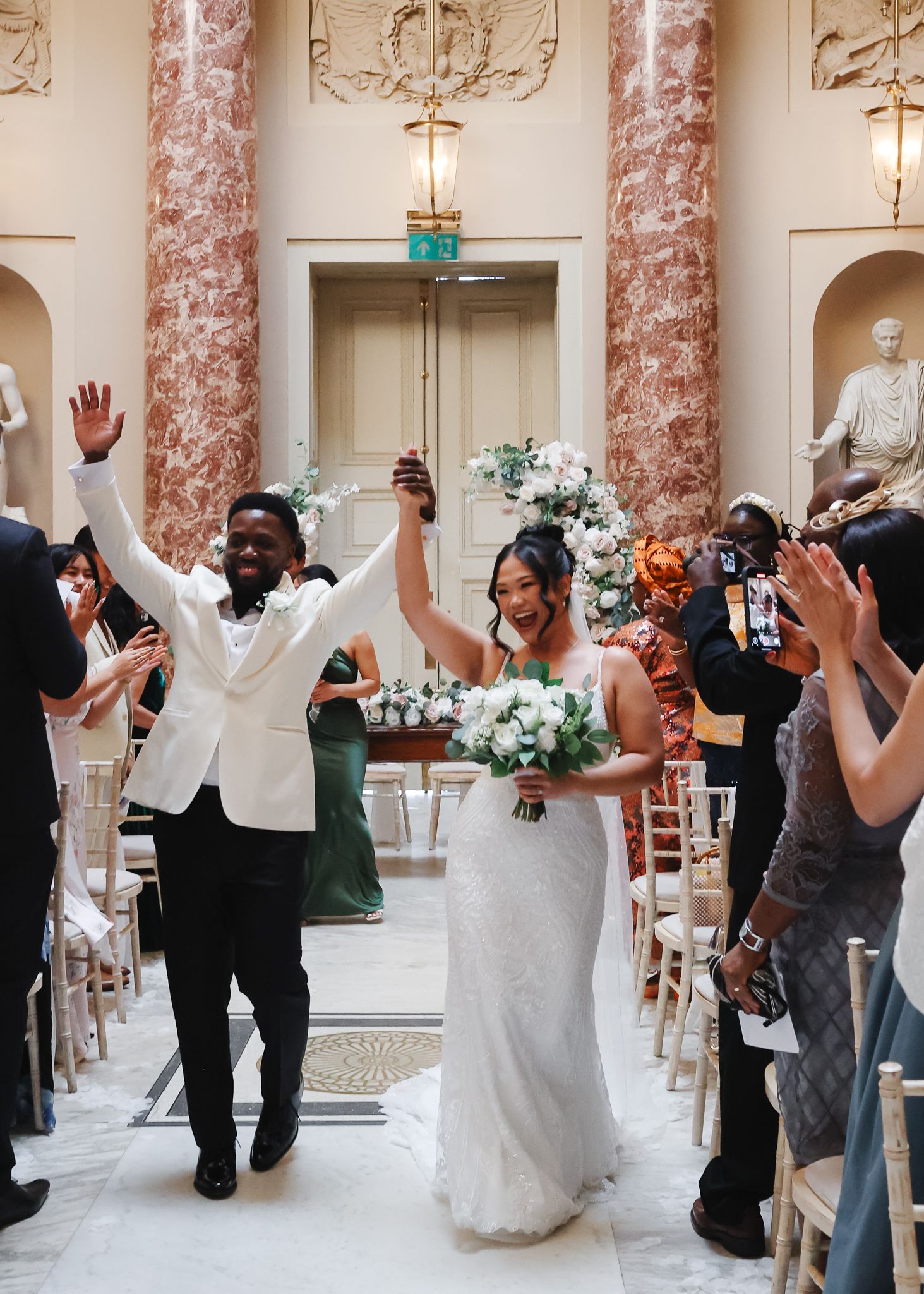 Wedding party celebrates with joyful dance moves in an elegant marble-columned venue decorated with white floral arrangements.