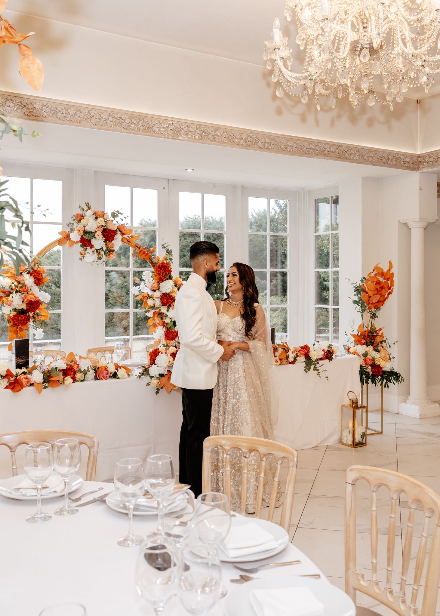 Elegant wedding reception setup with orange floral arch, crystal chandelier, and formal place settings in white room.