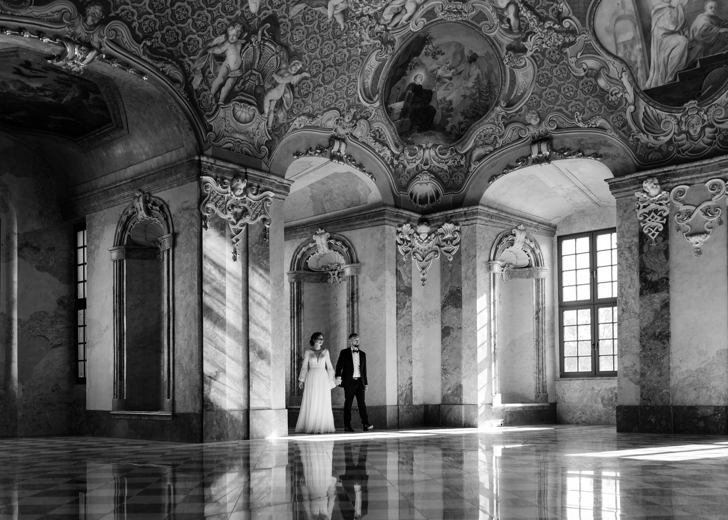 A black and white wedding photo in a grand baroque palace room with ornate ceiling frescoes and marble floors.