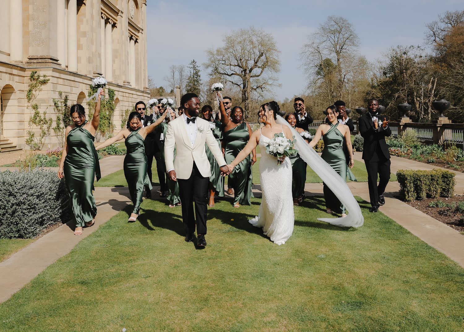 Wedding party stands in line on a grassy lawn outside a historic stone building during a spring celebration.