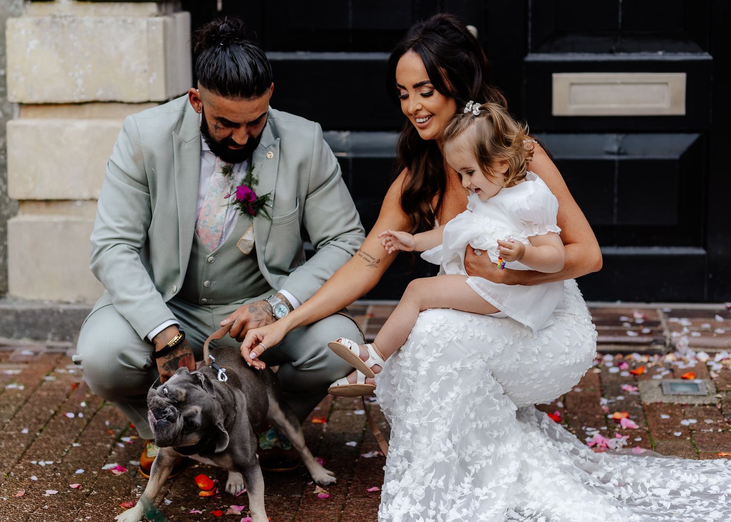 A couple in wedding attire pets a gray dog while sitting on steps surrounded by rose petals.