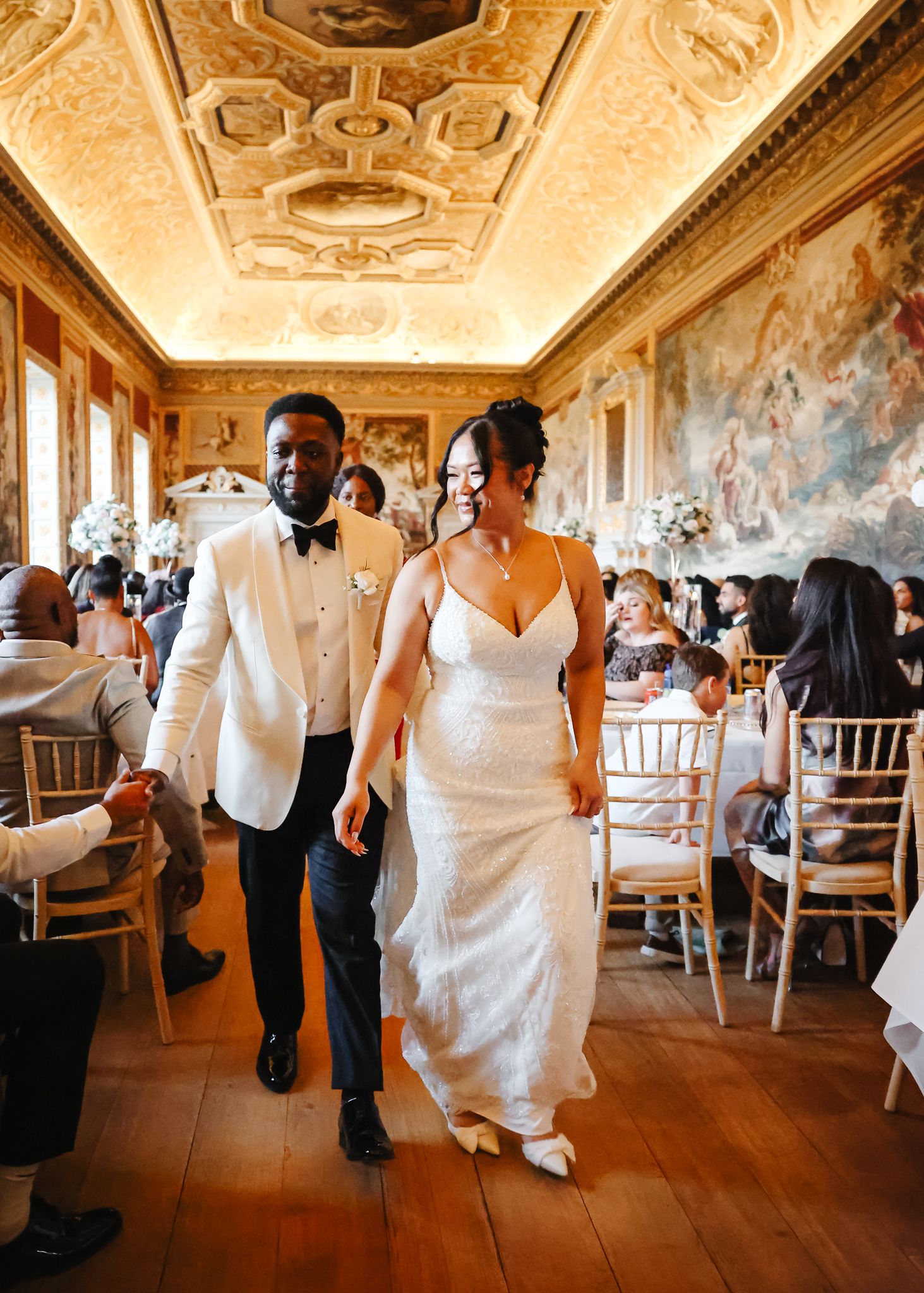 Guests dance and celebrate at an elegant wedding reception in an ornate ballroom with decorative ceiling and hardwood floors.
