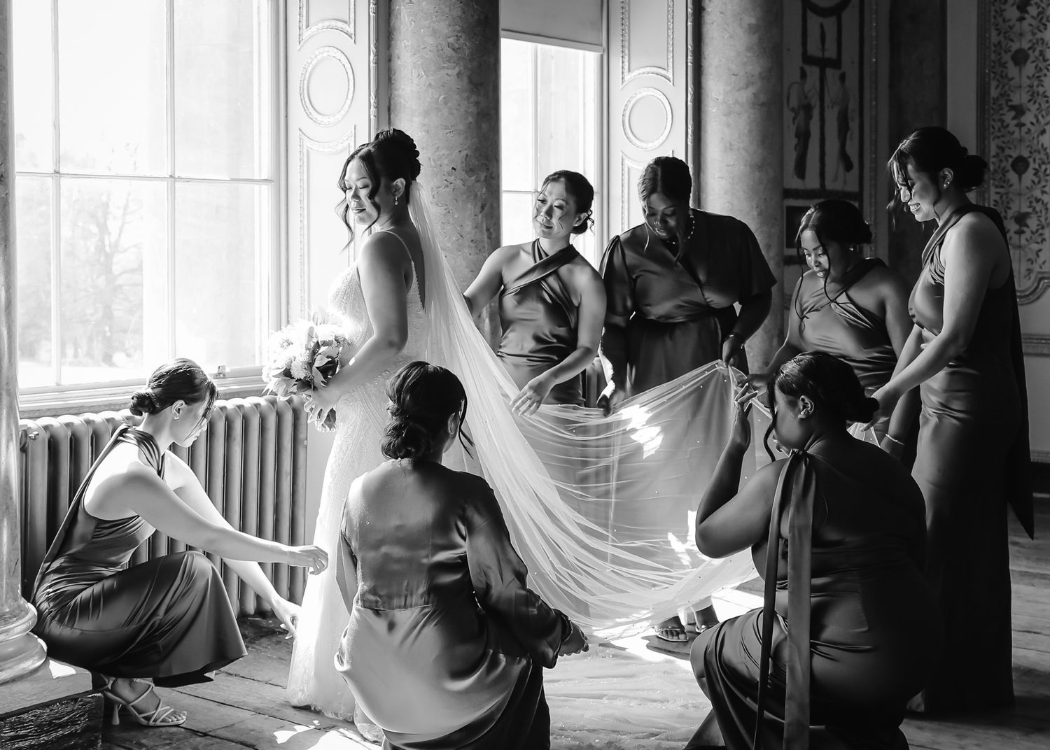 Wedding party helps with dress preparations in a sunlit room with classical columns in black and white photography.