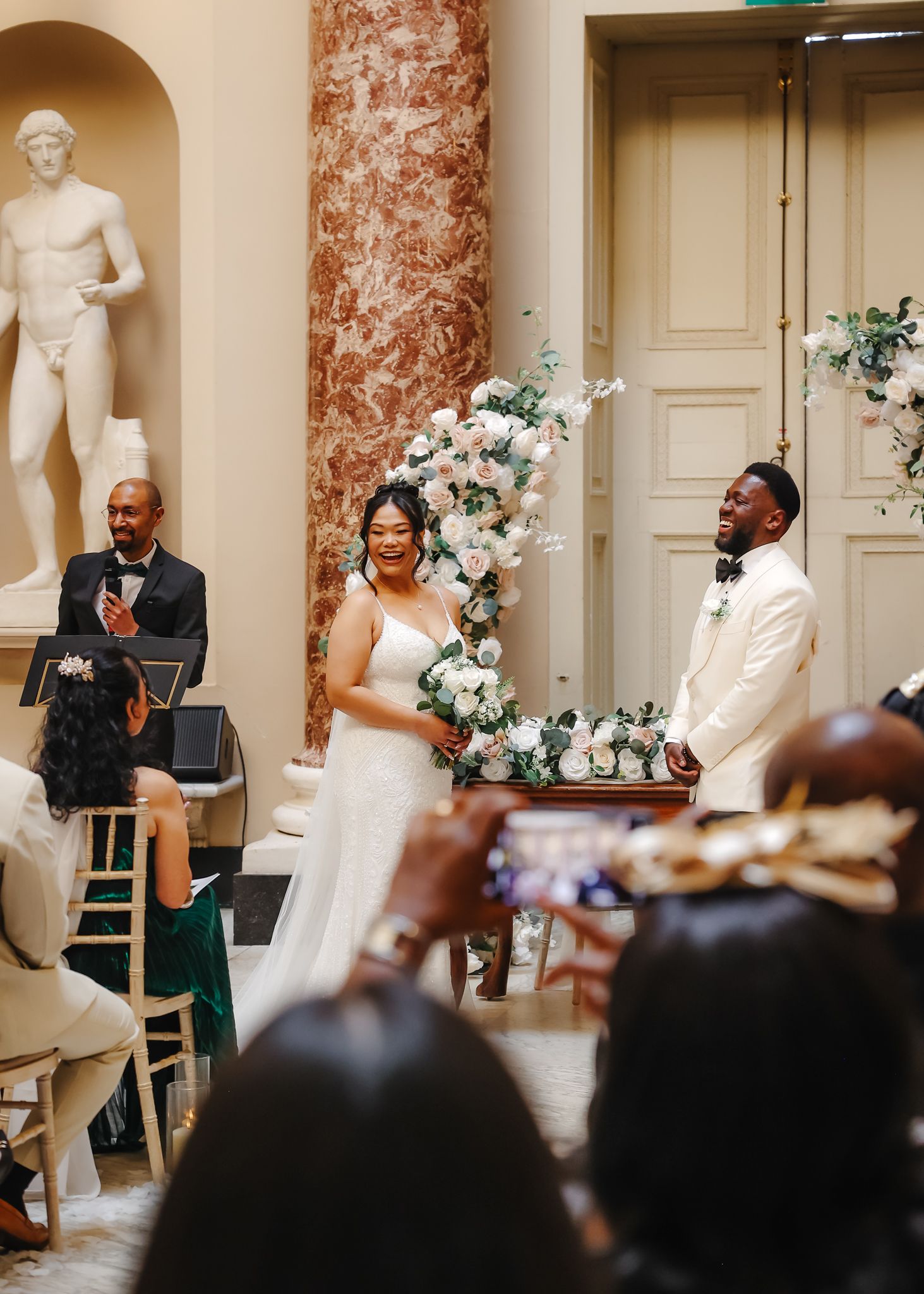 Wedding ceremony in an elegant marble hall with classical statues and floral decorations along the archways.