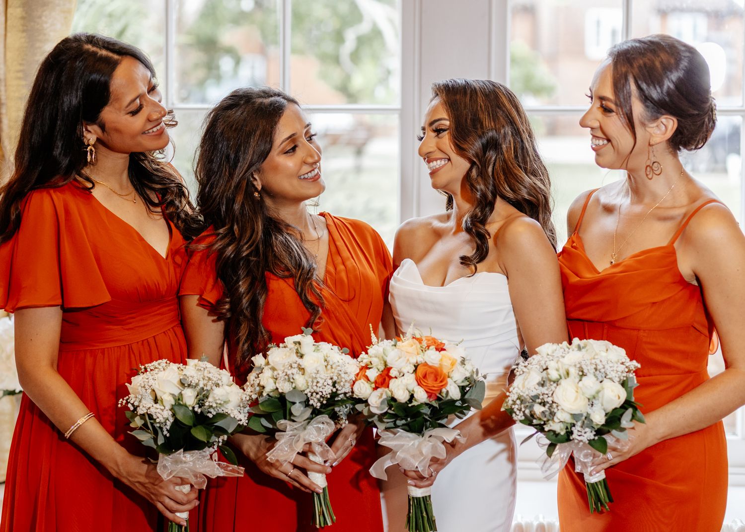 A bridal party wearing vibrant orange dresses holds white and orange floral bouquets while sharing a joyful moment together.