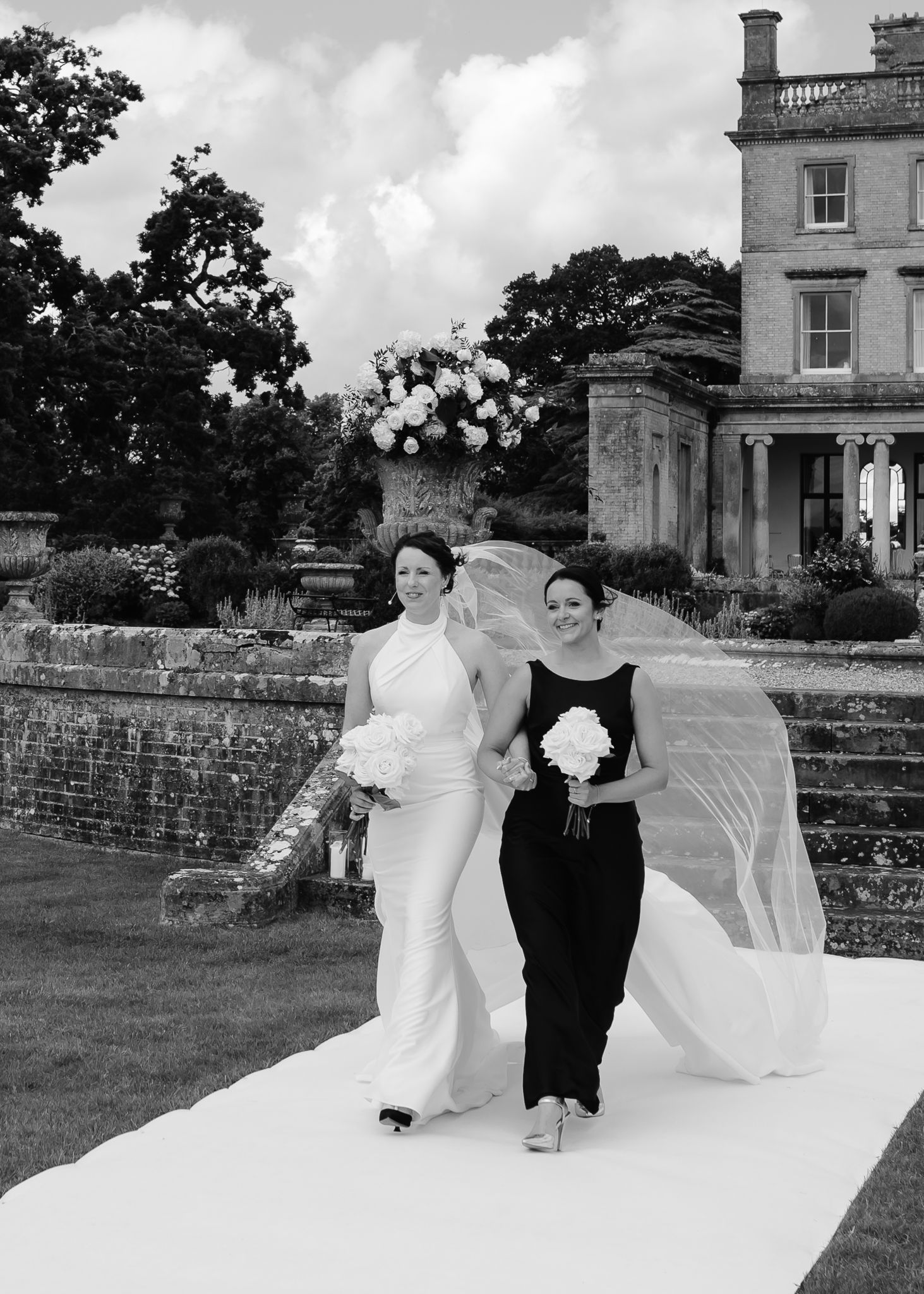 Two people walk down a white aisle runner at an elegant estate wedding with floral arrangements.
