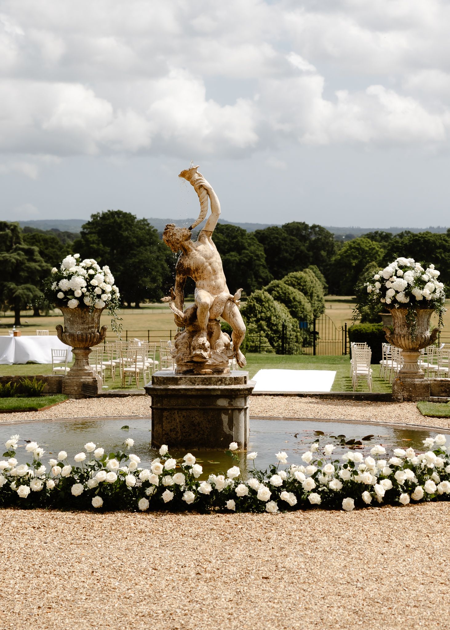 Elegant garden wedding setup with white flowers and classical stone statues along a gravel path.
