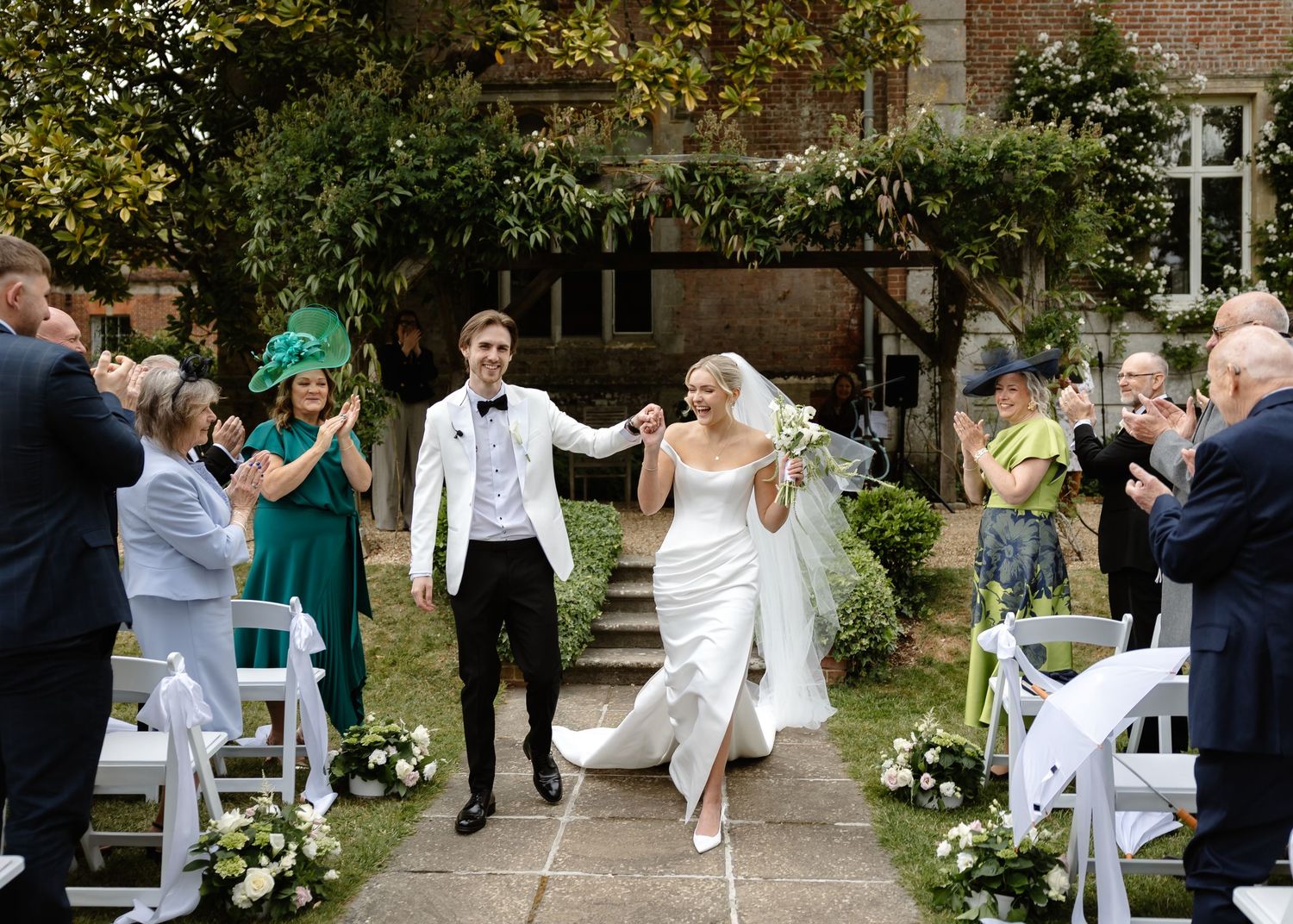 Bride and groom walk hand in hand down outdoor aisle, guests applauding, lush garden setting at elegant brick venue.