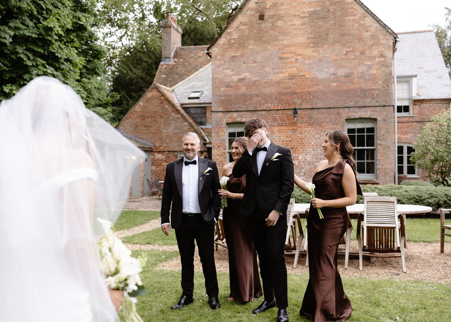 Wedding party in formal attire laughing outdoors near a brick manor house, with bride's veil visible in foreground.