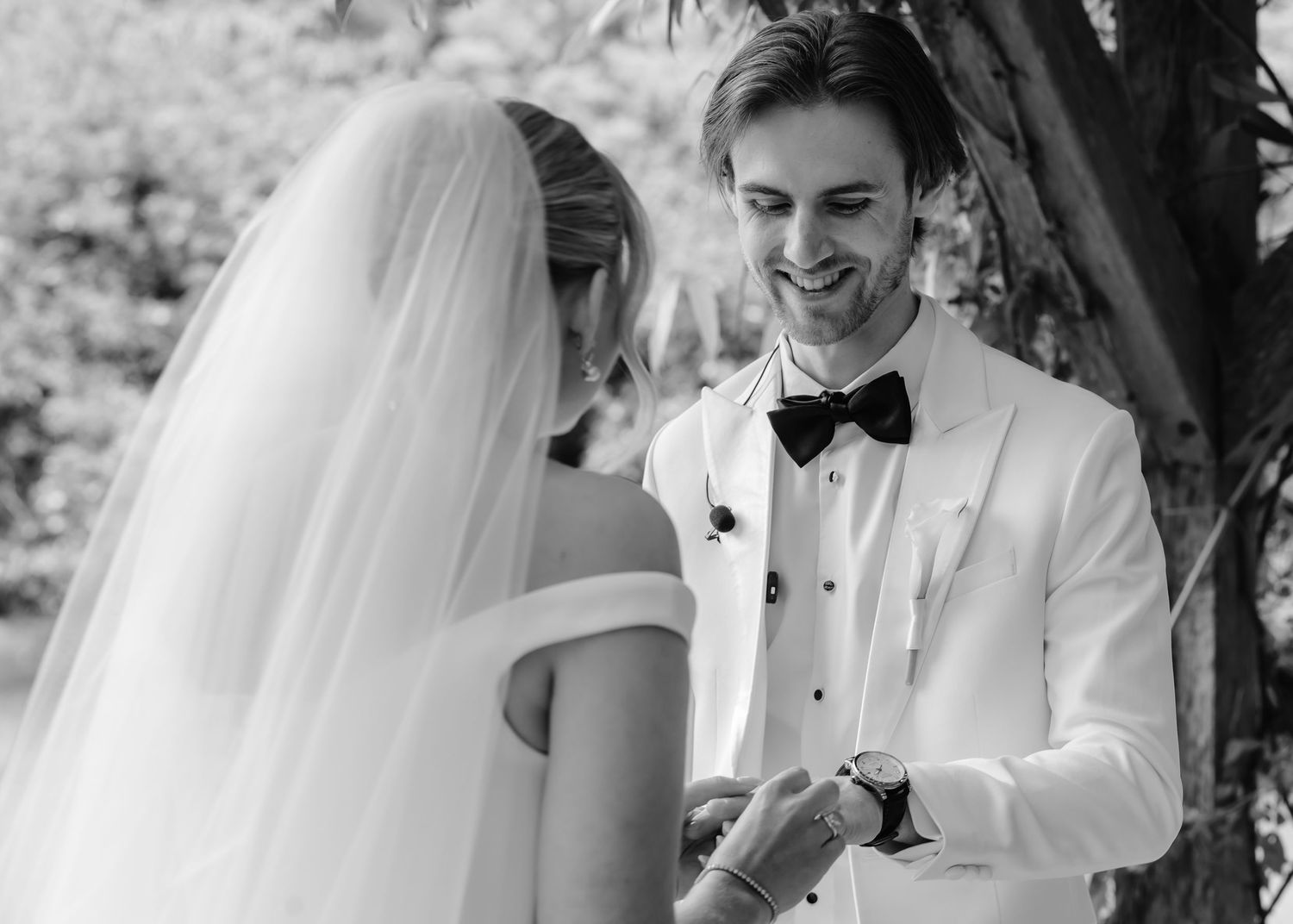 Black and white photo of smiling groom in white tuxedo placing ring on bride's finger during outdoor wedding ceremony.