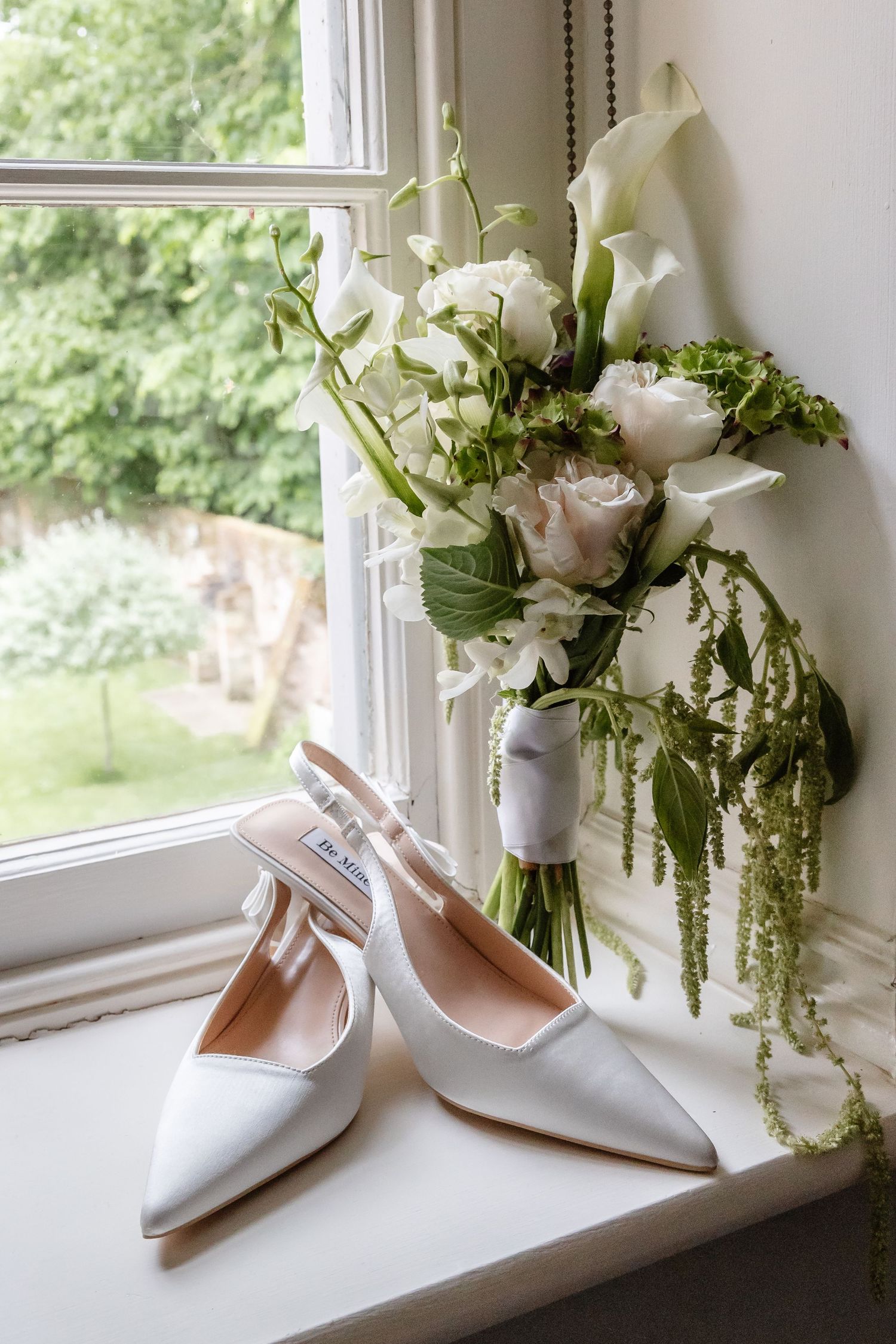 White slingback bridal heels on windowsill beside elegant bouquet of calla lilies and blush roses with greenery.