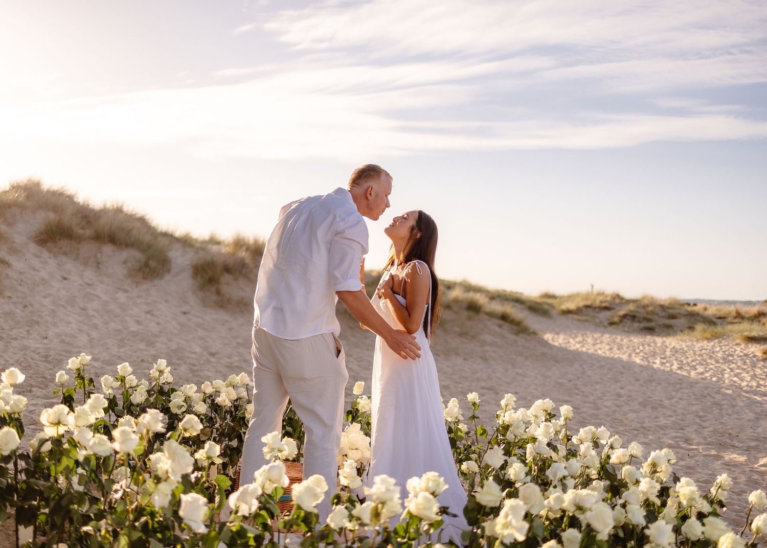 Couple sharing a kiss at sunset on a sandy beach surrounded by white roses in a romantic outdoor ceremony.