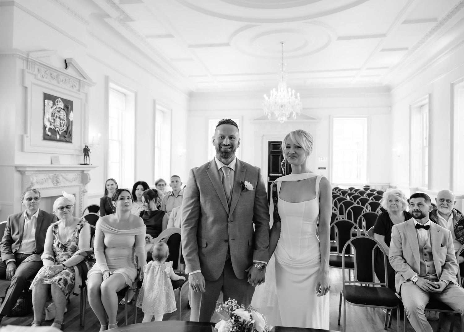 Bride and groom stand together at wedding ceremony altar in elegant white hall with seated guests.