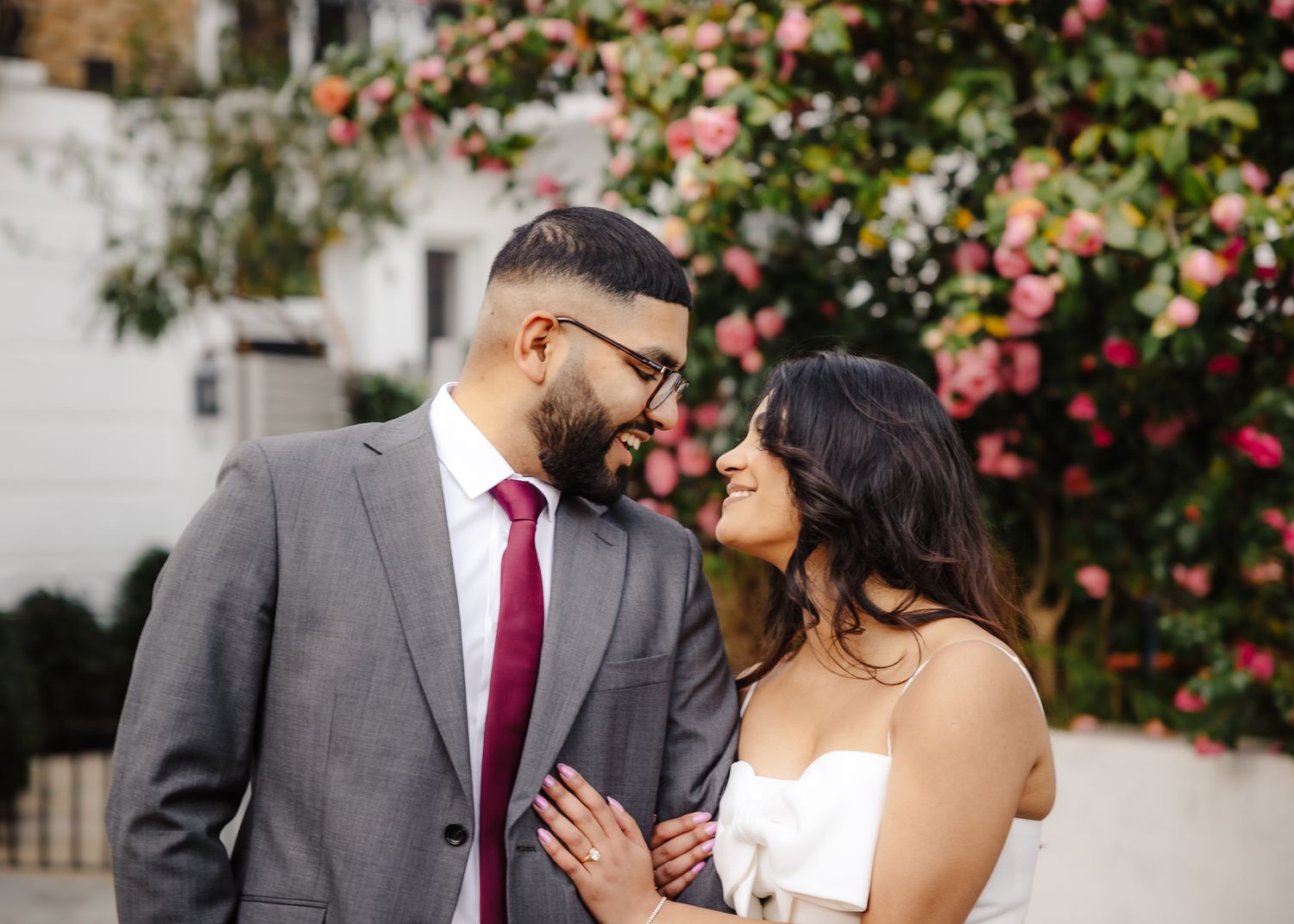 A couple in wedding attire smiles at each other in front of pink flowering bushes, groom in gray suit with burgundy tie.