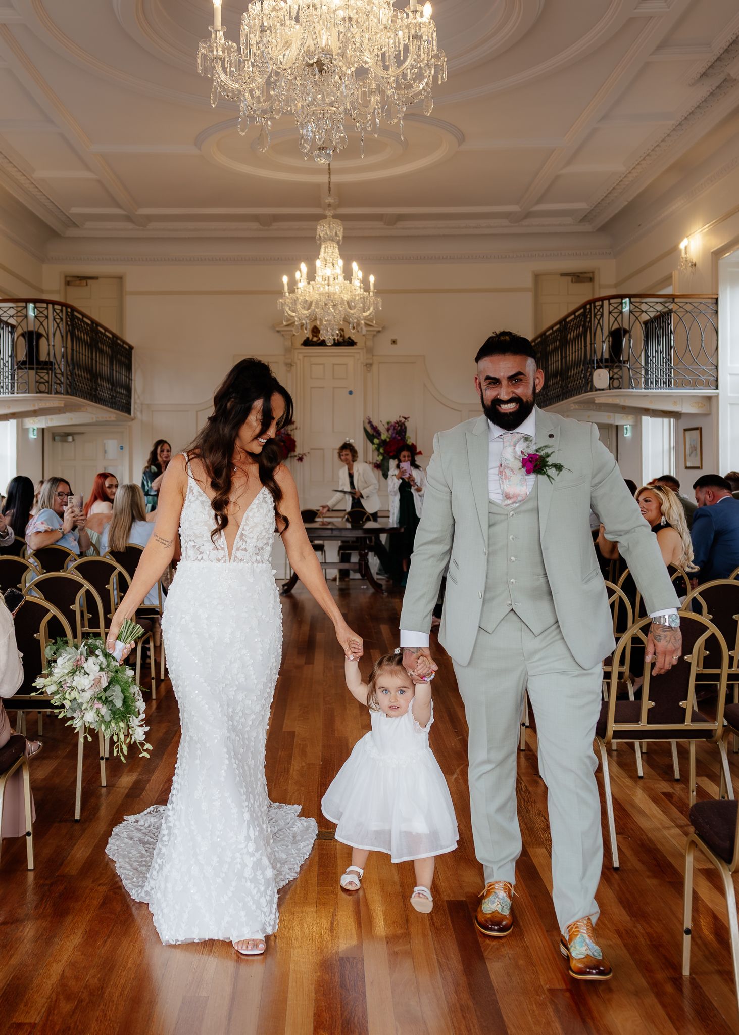 Wedding party dancing and celebrating in grand ballroom with ornate chandeliers and hardwood floors.