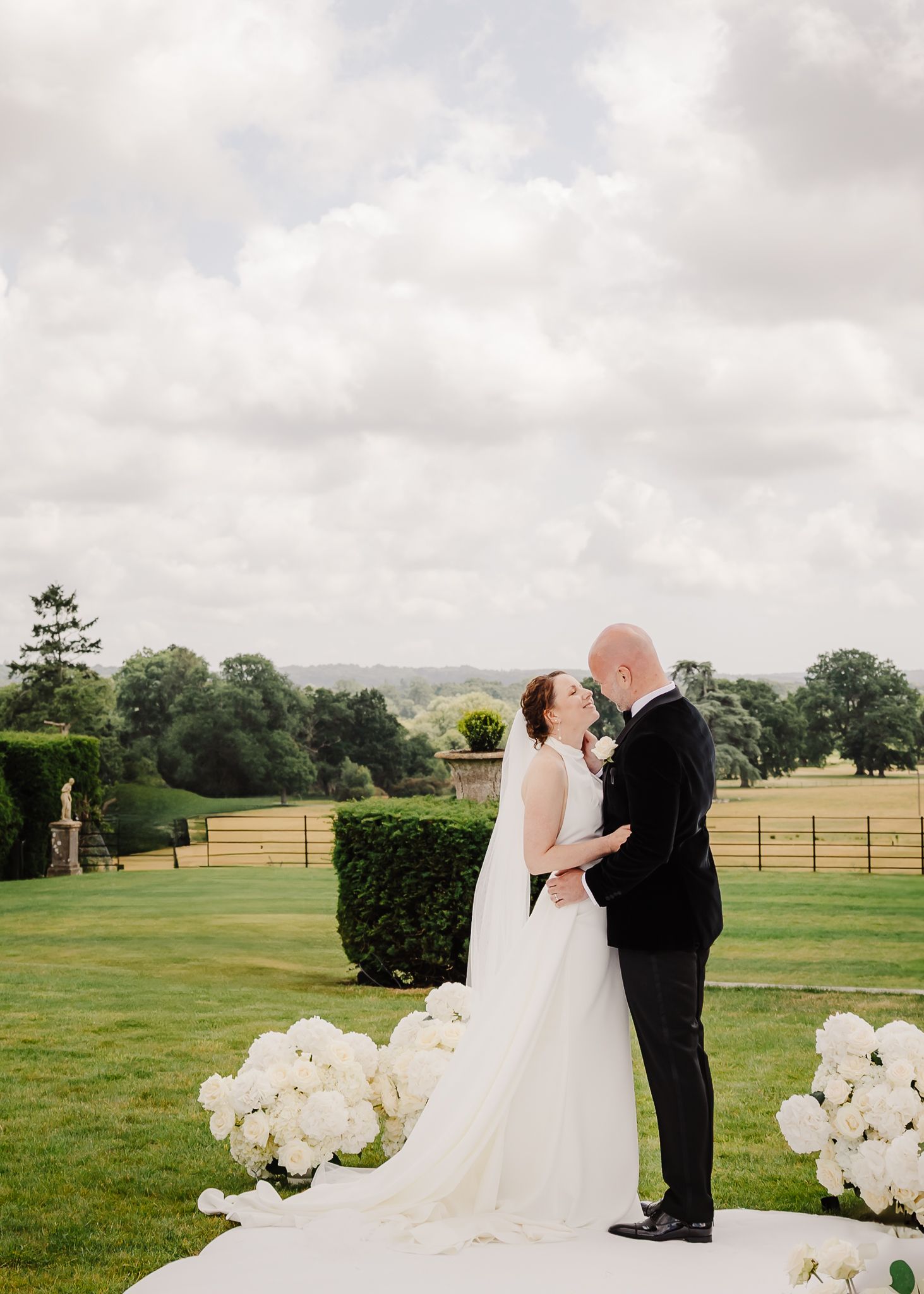 A couple shares a romantic moment on their wedding day in a manicured garden with white floral decorations.