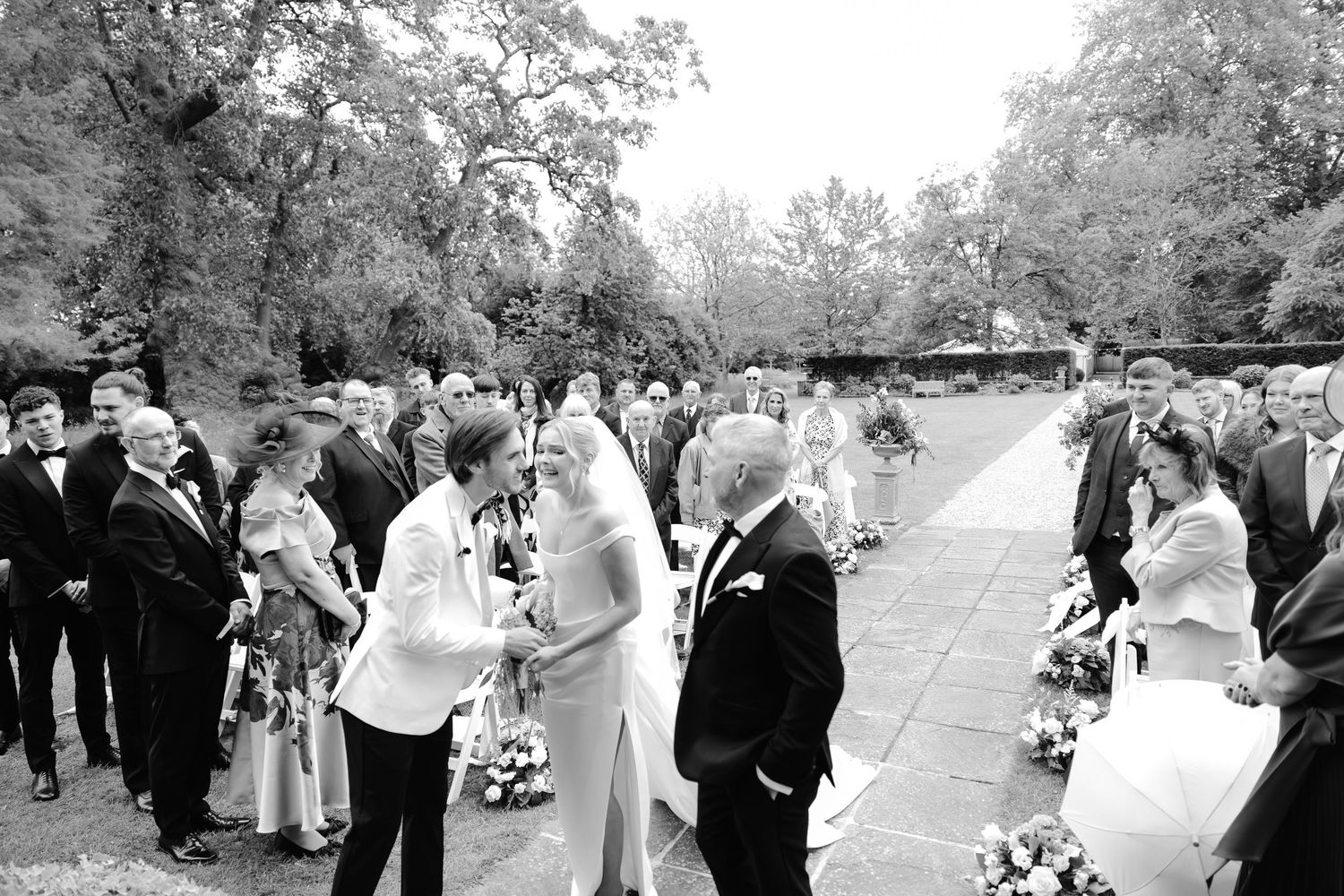 Black and white outdoor wedding ceremony with guests gathered on a patio surrounded by lush trees.