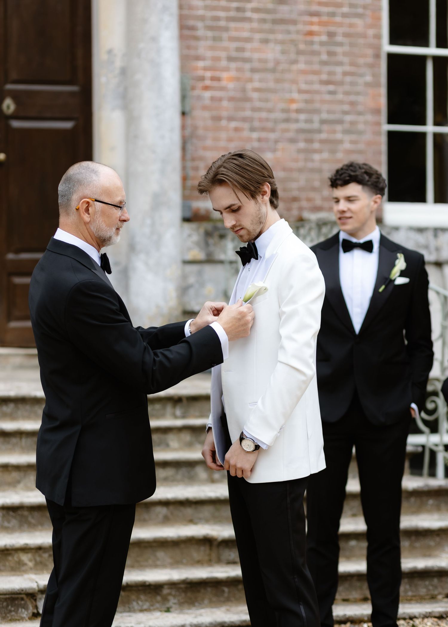 Older man pinning boutonniere on groom in white tuxedo jacket before wedding ceremony on stone steps.