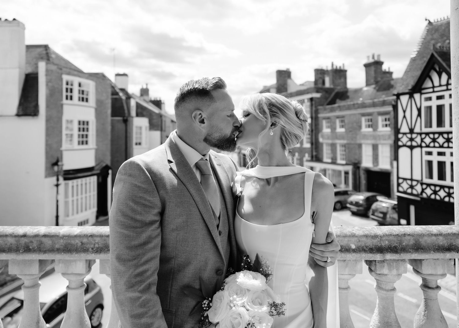 Bride and groom sharing a kiss on balcony with charming town buildings in black and white photo.