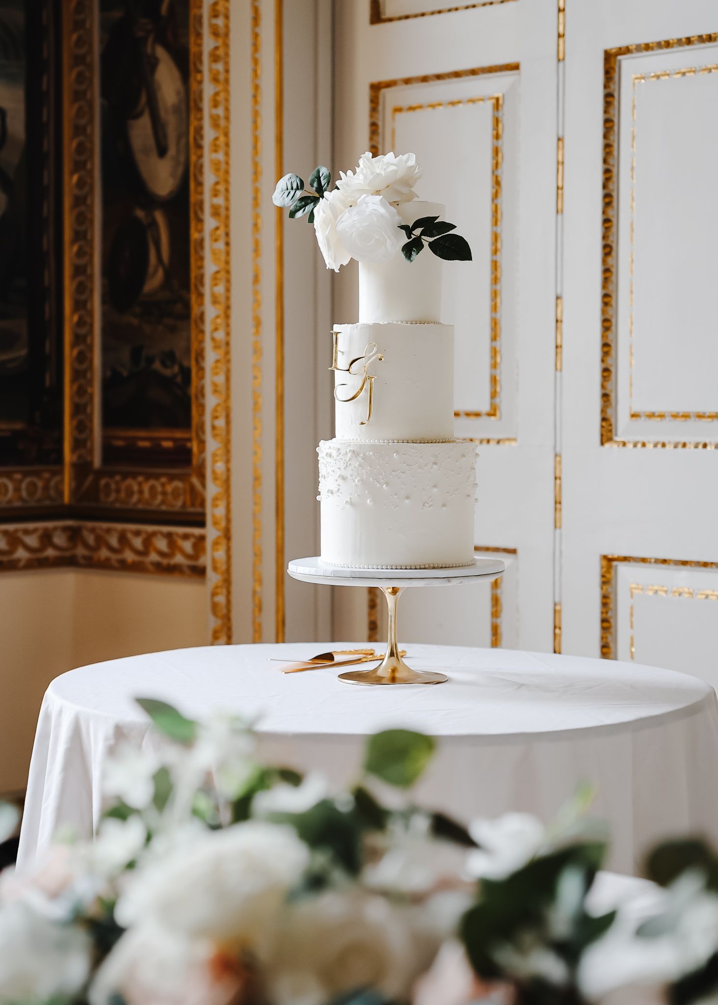 White tiered wedding cake adorned with white roses and greenery against a classic paneled wall with gold trim.
