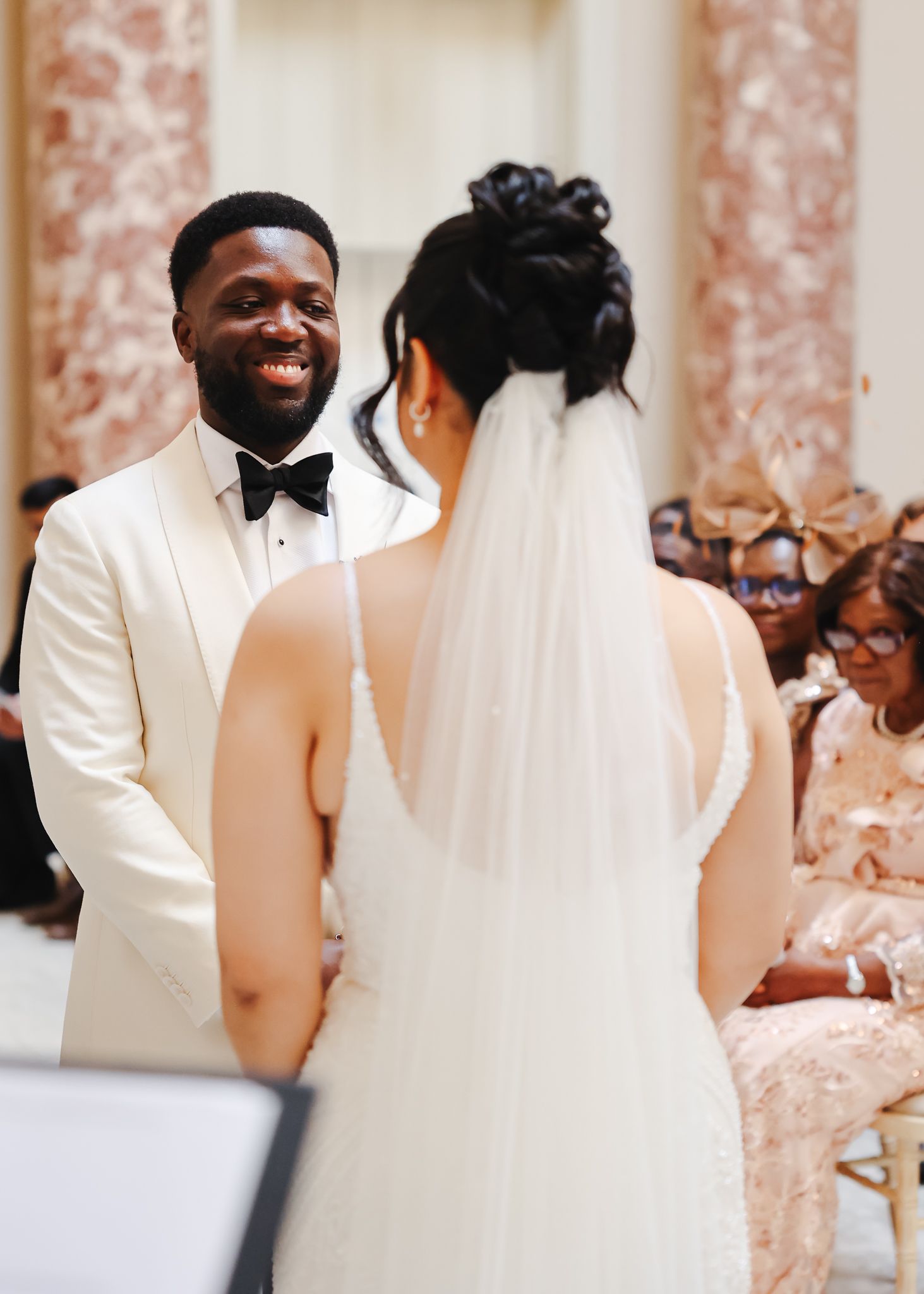 A wedding party wearing white tuxedos and gowns pose together in an elegant venue with floral decor.
