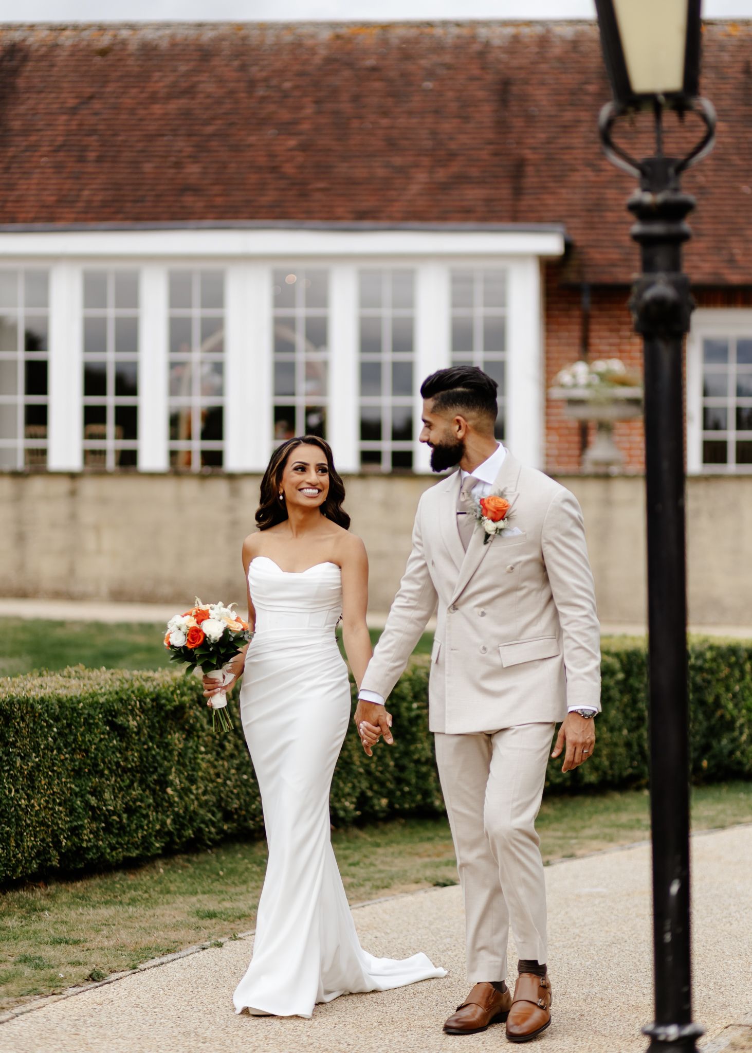 A couple in elegant wedding attire walk together on a garden path, with the bride in white and groom in beige suit.