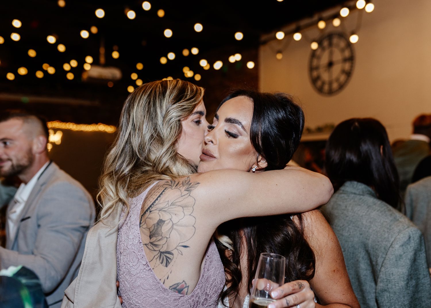 Two guests share an emotional embrace at an indoor wedding reception with twinkle lights overhead.