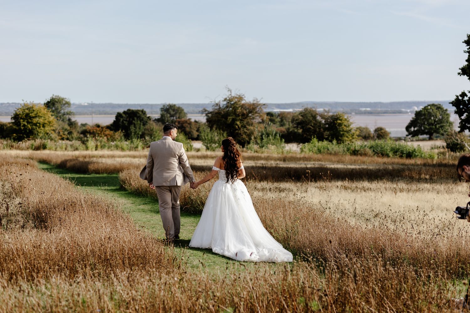 A couple walks through a scenic field of tall grass near the water on their wedding day.