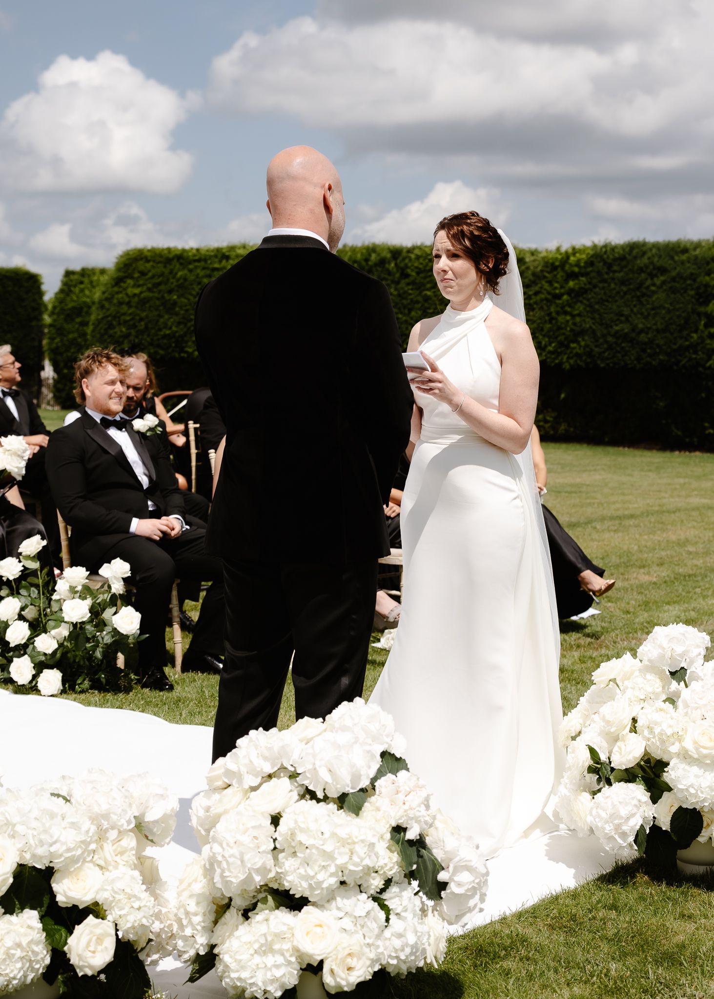 Wedding ceremony on a lush lawn with abundant white flowers lining the aisle.