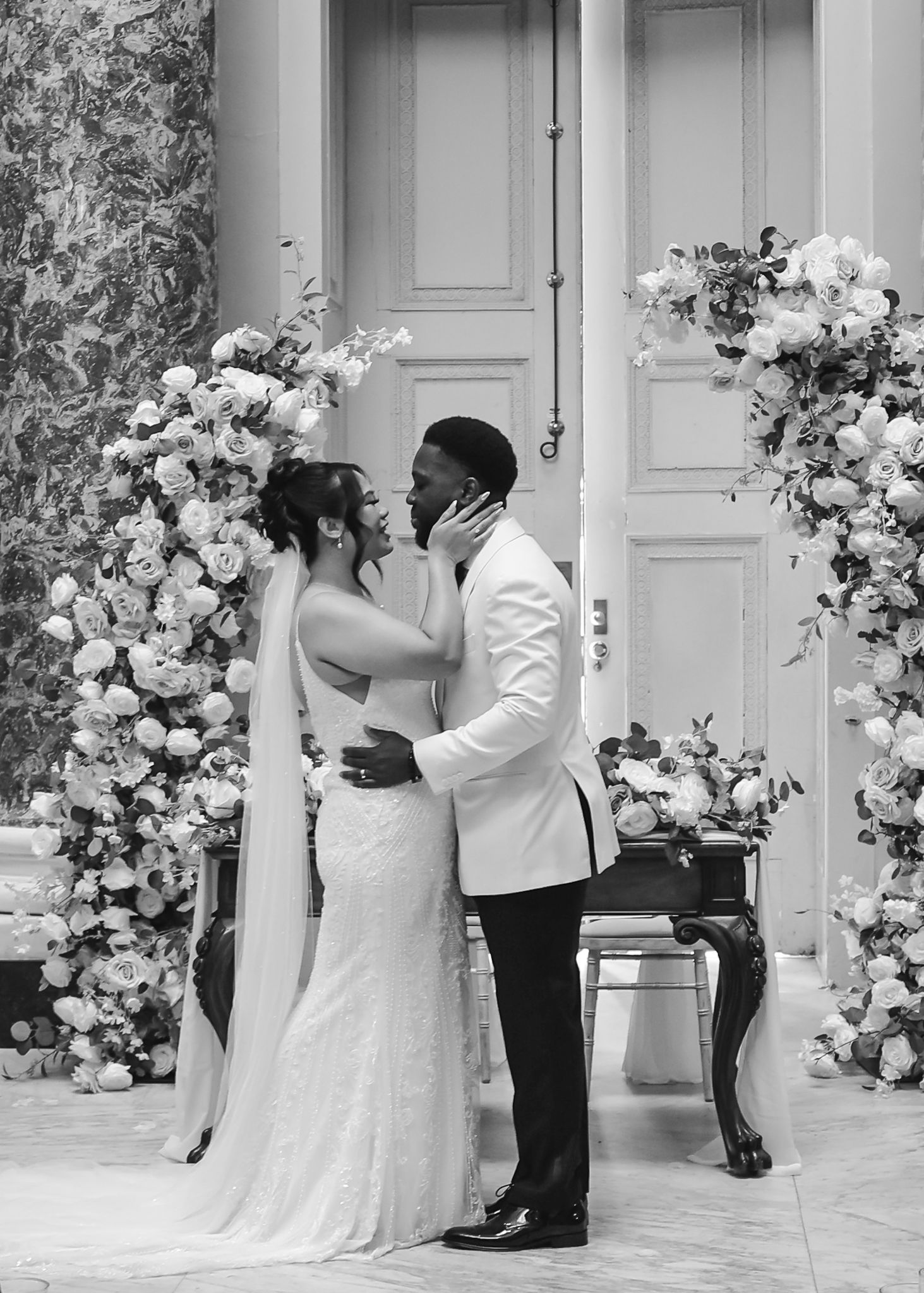 Romantic wedding couple shares intimate first kiss moments in front of elegant white door with floral decorations.